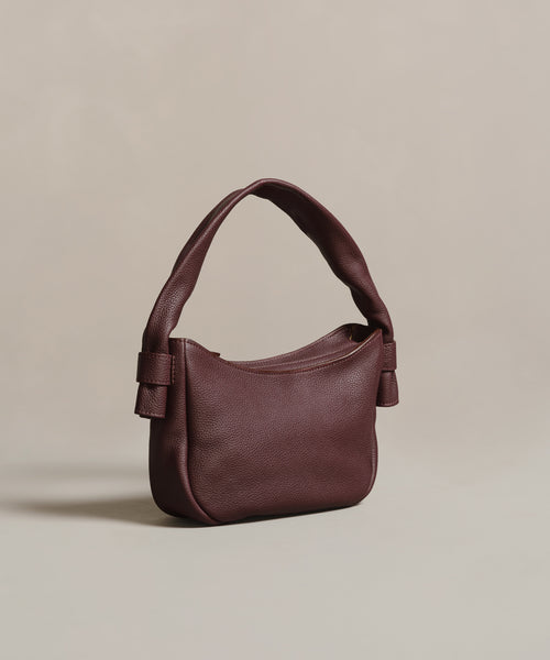 The Loren Shoulder Bag, crafted from soft dark brown leather with a curved padded handle and subtle side knots, is expertly made in Italy and displayed against a neutral beige background.