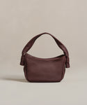 The Loren Shoulder Bag, a small Italian-made accessory, features dark brown soft leather, a curved handle, and knot details on each side, shown against a plain beige background.