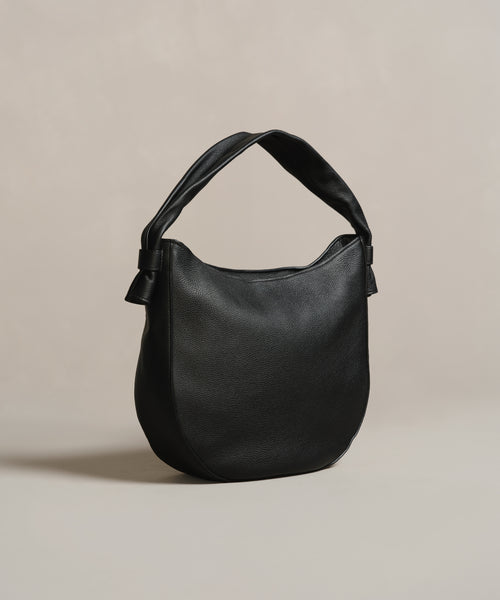 The Loren Tote is an Italian-made black leather shoulder bag featuring a wide padded strap and minimalist rounded design, shown against a neutral beige background.
