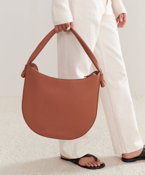 A person in white pants, a cream sweater, and black sandals holds the Loren Tote—a large, crescent-shaped Italian leather bag with a thick handle. The look is minimal and stylish.