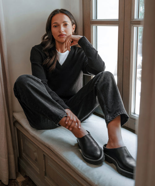 A woman with long brown hair sits on a window seat, wearing black jeans and the Leather Lug Mule. With one arm resting on her knee, she gazes calmly at the camera as natural light streams through the window.