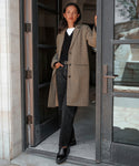 A woman with straight dark hair stands confidently in a doorway, wearing chic black Leather Lug Mules with a long brown coat, black top, and black jeans, one hand resting on the doorframe as she looks at the camera.