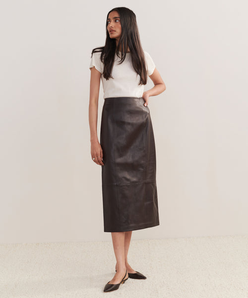 A woman stands against a plain background wearing a white short-sleeve top, the high-waisted Leather Sienna Skirt in chocolate, and pointed brown flats. She looks to the side with one hand on her hip.