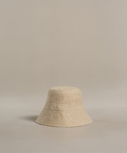 The Linen Bucket Hat, crafted in Portugal from beige linen, features a simple design and downward-sloping brim. Photographed on a neutral background, it’s the perfect minimalist accessory for summer beach days.