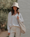 A woman stands outdoors in a beige Linen Bucket Hat with a matching shoulder bag, loose white sweater, and light pants. Greenery and a white wall blur in the background.