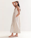 Woman with long dark hair models the Cove Dress, a sleeveless style with side pockets and a relaxed fit in Natural/Navy Stripe. She stands sideways in flat sandals against a plain light background.