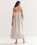 A woman with long dark hair stands barefoot on a light floor, wearing the Cove Dress. This sleeveless, ankle-length dress has thin straps, vertical stripes in Natural/Navy, and offers a relaxed fit as she faces away.