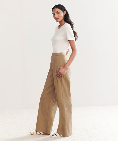 A woman with long, wavy dark hair and a neutral expression stands sideways in a minimal, light-colored room wearing a white short-sleeve top and the high-waisted khaki Linen Keaton Pant, styled with white flat sandals.