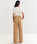 A woman with long dark hair, seen from behind, wears a white short-sleeve tee tucked into high-waisted, wide-leg Linen Keaton Pants in khaki and light-colored shoes, standing against a plain light background.