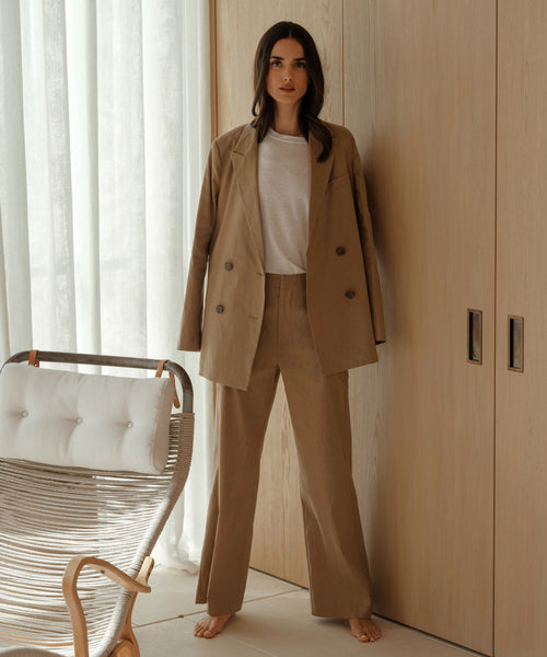 A woman stands barefoot indoors in the Linen Keaton Blazer over a white t-shirt, beside a modern wooden chair and light wood-paneled wall, with sheer curtains letting in natural light.