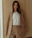 A woman with long dark hair stands confidently indoors against a wooden wall, wearing the Linen Keaton Blazer—a beige double-breasted style—over a white t-shirt.