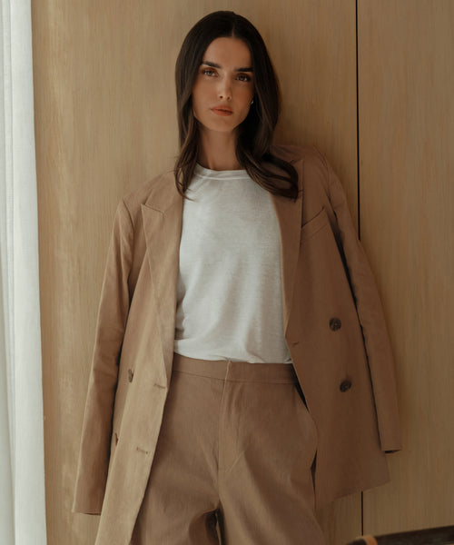 A woman with long dark hair stands confidently indoors against a wooden wall, wearing the Linen Keaton Blazer—a beige double-breasted style—over a white t-shirt.
