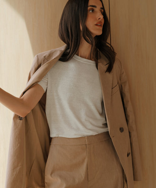 A woman with long brown hair wears the Linen Keaton Blazer with matching pants over a white t-shirt, standing against a light wooden background where sunlight casts soft shadows.