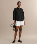 A woman stands against a beige background wearing the black Linen Logan Shirt, white shorts, black woven slip-on shoes, and holding a small black and straw handbag.