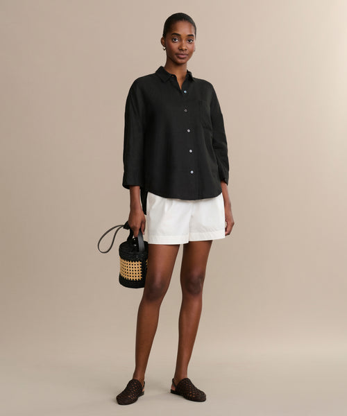 A woman stands against a beige background wearing the black Linen Logan Shirt, white shorts, black woven slip-on shoes, and holding a small black and straw handbag.