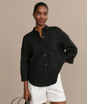 A woman stands against a beige background, wearing the black Linen Logan Shirt with long sleeves and white shorts. She holds a black bag in one hand, smiling softly with her hair pulled back.