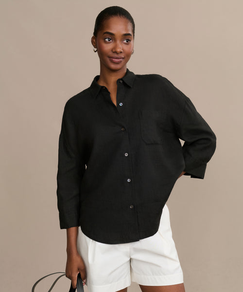 A woman stands against a beige background, wearing the black Linen Logan Shirt with long sleeves and white shorts. She holds a black bag in one hand, smiling softly with her hair pulled back.