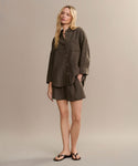 A woman poses in the boxy-fit Linen Logan Shirt with matching olive green shorts and black sandals, showcasing effortless style against a plain beige background.
