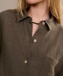 A person with long blonde hair wears the olive-green Linen Logan Shirt—boxy fit, chest pocket—paired with a short black beaded necklace for an effortless style. The face is partially out of frame.
