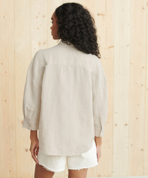 A woman with curly hair, seen from behind, wears the Linen Logan Shirt, a loose, boxy-fit button-up made from 100% linen, and white frayed shorts while standing in front of a light wooden wall.