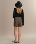 A person with long blonde hair, in a black hat, black long-sleeve top, Linen Logan Short in dark moss, and black sandals stands facing away against a beige background—showcasing the warm-weather style of the Linen Logan Short.