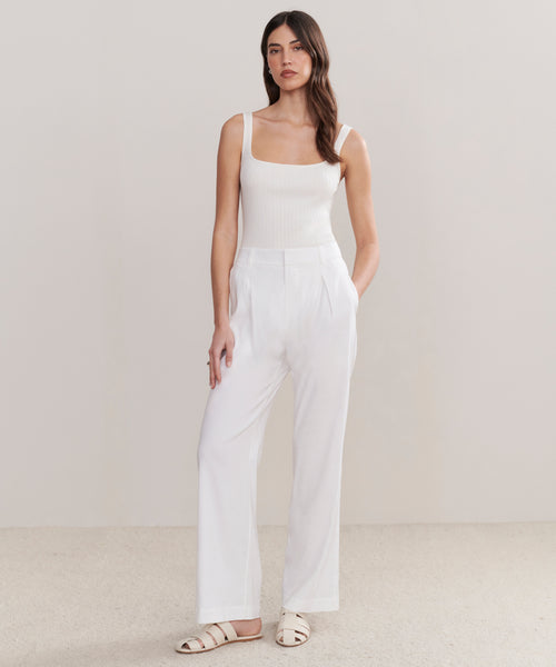 A woman with long, wavy brown hair stands against a plain background wearing the Riley Trouser—relaxed fit, high-waisted linen trousers—with a white ribbed tank top and white sandals. Her hands are in her pockets.