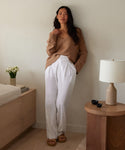 A woman with long dark hair, a neutral expression, and sandals stands indoors by a wooden table, wearing the Riley Trouser and a tan sweater. Nearby is a bed with books and white flowers in a vase.