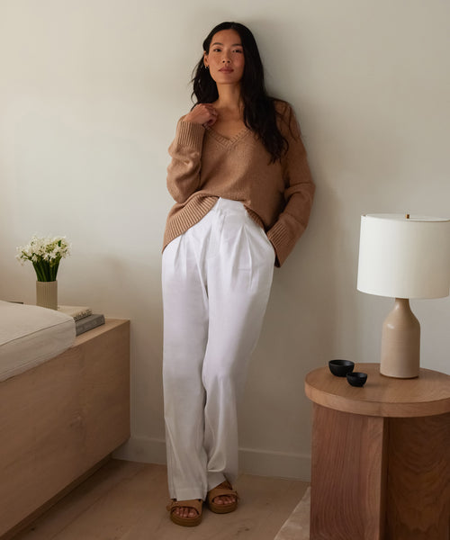 A woman with long dark hair, a neutral expression, and sandals stands indoors by a wooden table, wearing the Riley Trouser and a tan sweater. Nearby is a bed with books and white flowers in a vase.