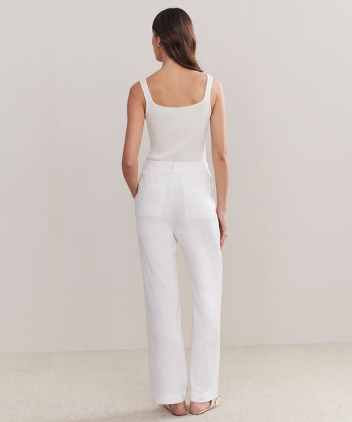 A woman faces away with her hands in her pockets, wearing a white sleeveless ribbed top and Riley Trouser high-waisted relaxed fit summer trousers against a plain light background.