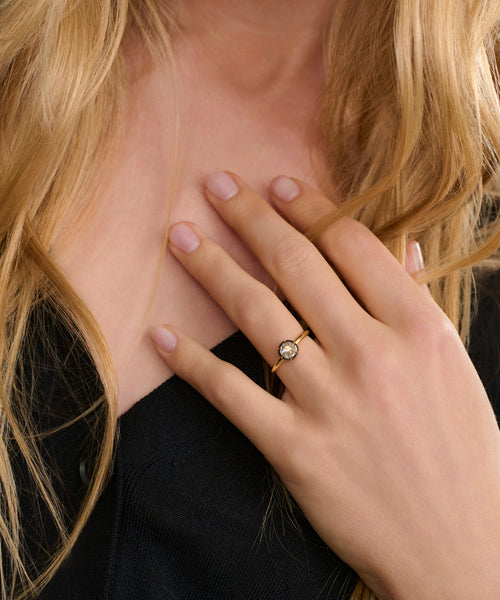 A person with long blonde hair in a black top touches their chest with their left hand, displaying the Liv Rose Cut Ring—a gold band with a round, clear gemstone—on their ring finger for a chic, modern accent to a classic style.
