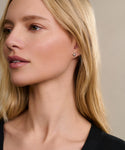 A woman with long blonde hair looks to the side, wearing a black top and the Liv Rose Cut Studs. Her skin is smooth, her makeup natural with soft pink lips, and the background is neutral.