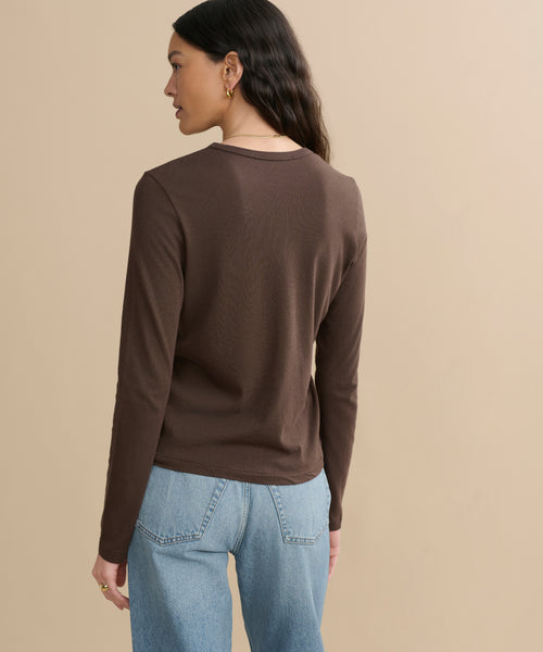 A woman with long dark hair stands with her back to the camera against a plain beige background, wearing the Long-Sleeve Slim Tee in brown and light blue jeans.