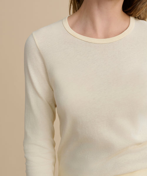 A person models the Long-Sleeve Slim Tee—a classic crewneck in soft, light cream 100% cotton—against a beige background, showing only their upper torso and part of their face.