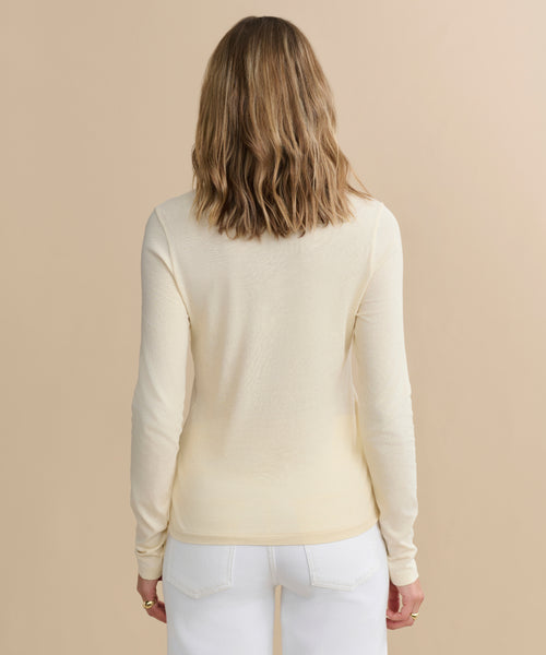 Seen from behind, a person with wavy, shoulder-length light brown hair wears a cream Long-Sleeve Slim Tee and white jeans, standing against a plain beige background.