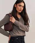 A woman with long dark hair in a gray sweater and jeans confidently holds the Loren Shoulder Bag over her shoulder, wearing gold earrings and a ring.