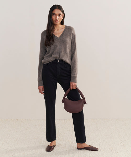 A woman with long dark hair holds the Loren Shoulder Bag in soft leather while wearing a taupe V-neck sweater, black high-waisted pants, and brown flats. She stands against a plain light background on a textured beige floor.