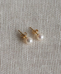 A pair of Small Loren Pearl Studs by Jenni Kayne Jewelry, featuring gold and round white pearls, rests on a textured off-white fabric background.