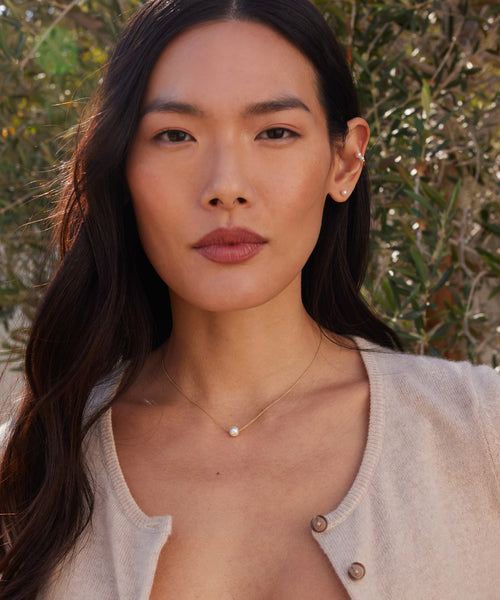 A woman with long dark hair wears a beige top, Jenni Kayne Jewelry—a delicate gold necklace and Small Loren Pearl Studs. She stands outdoors in natural light with greenery behind her.