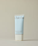 A light blue tube of Oak Ess Luminous Body Lotion Travel with Blue Tansy and a white cap stands upright on a beige background. The label shows minimalist text in soft brown and blue shades.