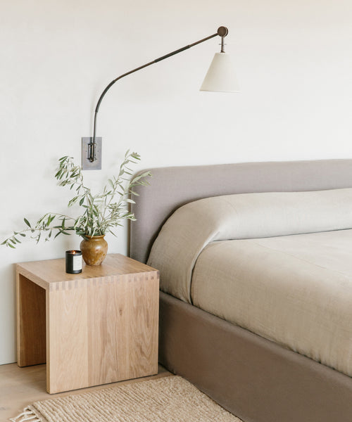 A lamp on a wall next to a bed is mounted above the Malibu Side Table, creating a clean and serene ambiance.