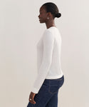 A woman, smiling and standing sideways against a plain light background, wears the white Mercer Crewneck with a classic cut, paired with dark blue jeans and hoop earrings. Her hair is styled in a neat low bun.