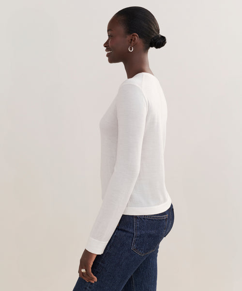 A woman, smiling and standing sideways against a plain light background, wears the white Mercer Crewneck with a classic cut, paired with dark blue jeans and hoop earrings. Her hair is styled in a neat low bun.