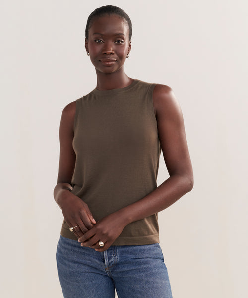 A woman with short hair stands against a light background, wearing the Merino Tank in olive green and blue jeans. She looks at the camera calmly, her hands gently clasped in front of her.