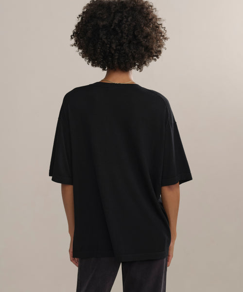 Someone with short curly hair, seen from behind, wears an oversized black Merino Tee with dark pants, standing before a plain light background.