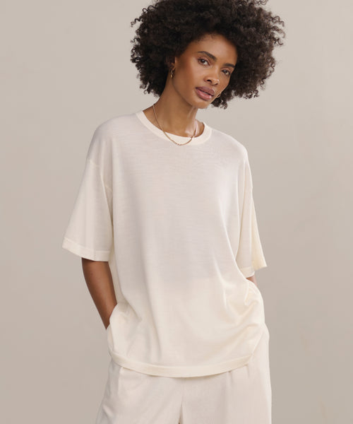 A woman with curly hair wears the Merino Tee in an oversized off-white style with matching pants, hands in pockets, standing against a plain background—an elevated basic for effortless style.