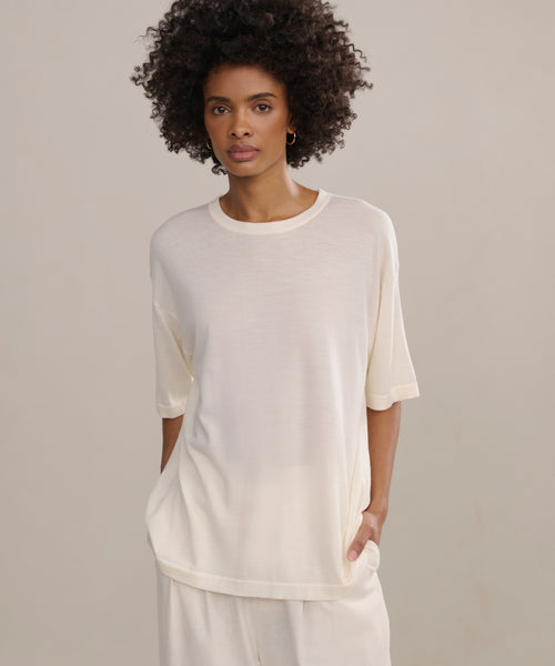 A person with short curly hair stands against a neutral background, wearing an oversized light cream Merino Tee and matching pants, one hand in their pocket.