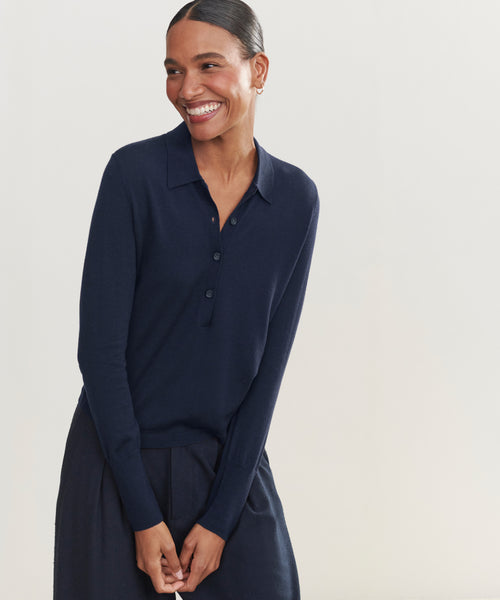 A dark-haired woman stands smiling against a plain light background, wearing the navy Grayson Pullover with matching bottoms and her hands clasped in front.