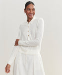 A smiling woman stands against a plain light background, wearing the Grayson Pullover—a cream-colored, casual polo-style sweater with buttons—paired with a matching skirt.