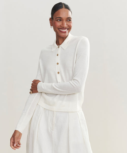 A smiling woman stands against a plain light background, wearing the Grayson Pullover—a cream-colored, casual polo-style sweater with buttons—paired with a matching skirt.