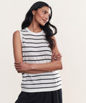 A woman with long, dark hair stands with arms crossed, wearing the Merino Tank in white with black stripes and black pants, looking to the side and smiling slightly against a plain light background.
